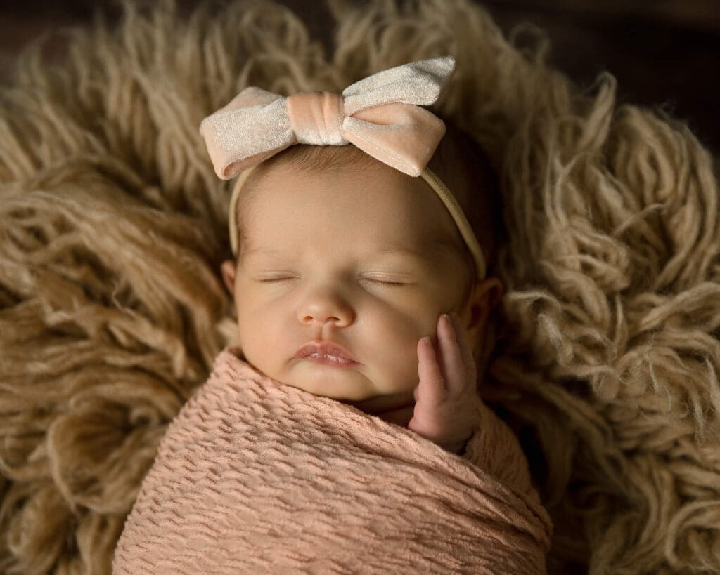 Newborn girl wrapped in a peach wrap and wearing a peach bow headband as she sleeps on a tan rug.