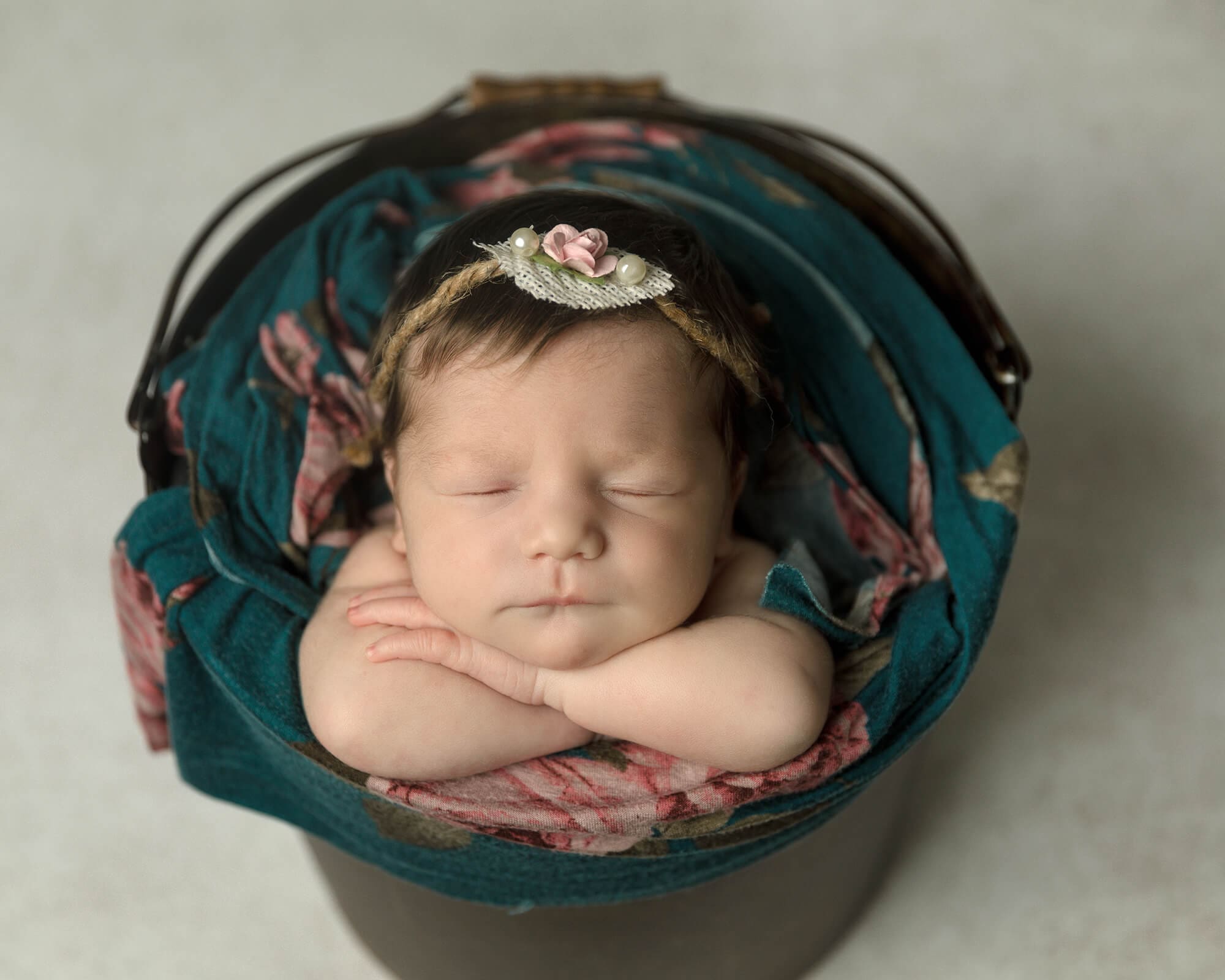 Newborn girl sleeping in a pail. Pail is layered with a teal floral wrap.