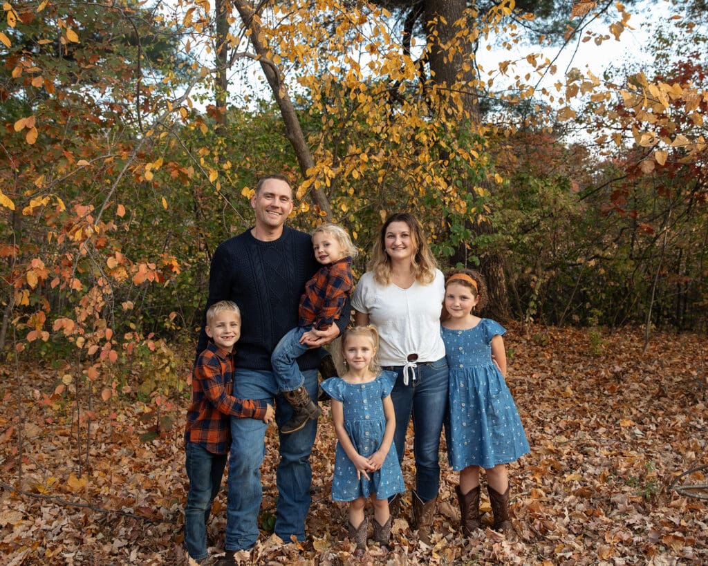 Family of 6 standing with fall colored trees in the background.