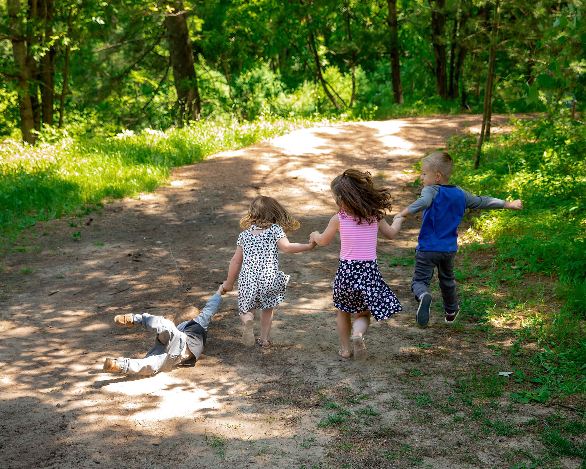 Four cousins holding hands running away into the woods. Youngest of cousins falls and is being dragged.