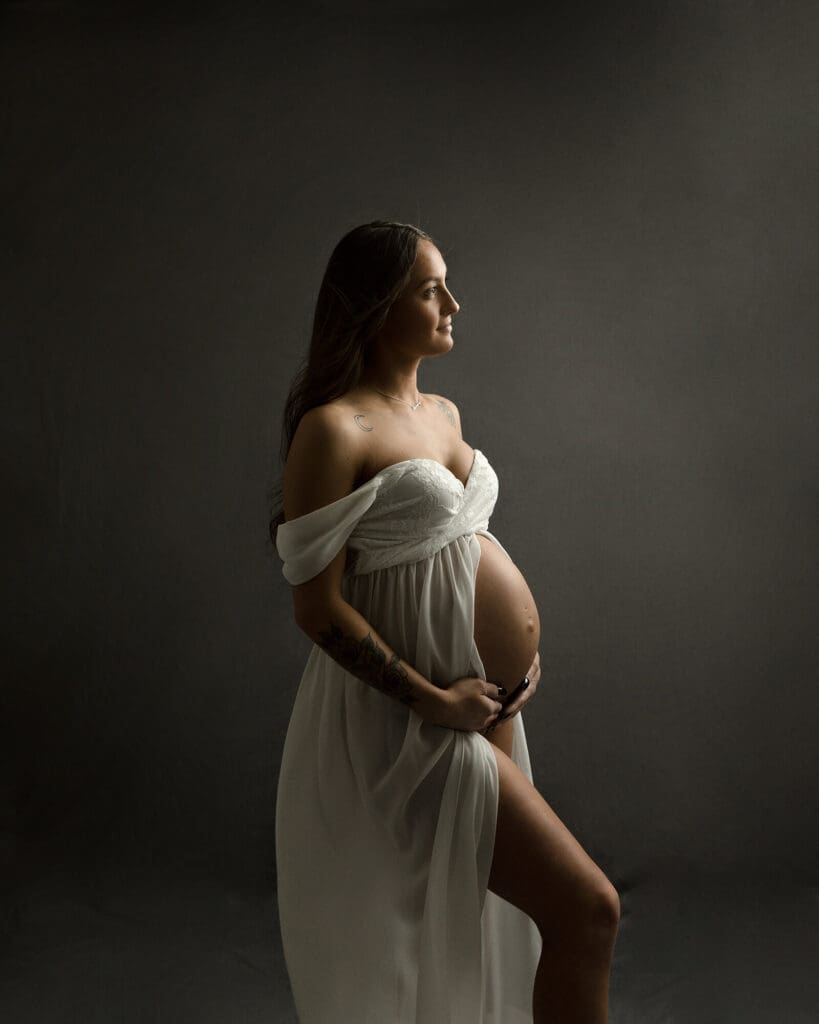 Pregnant woman in a white maternity gown backlit to show off her baby bump.