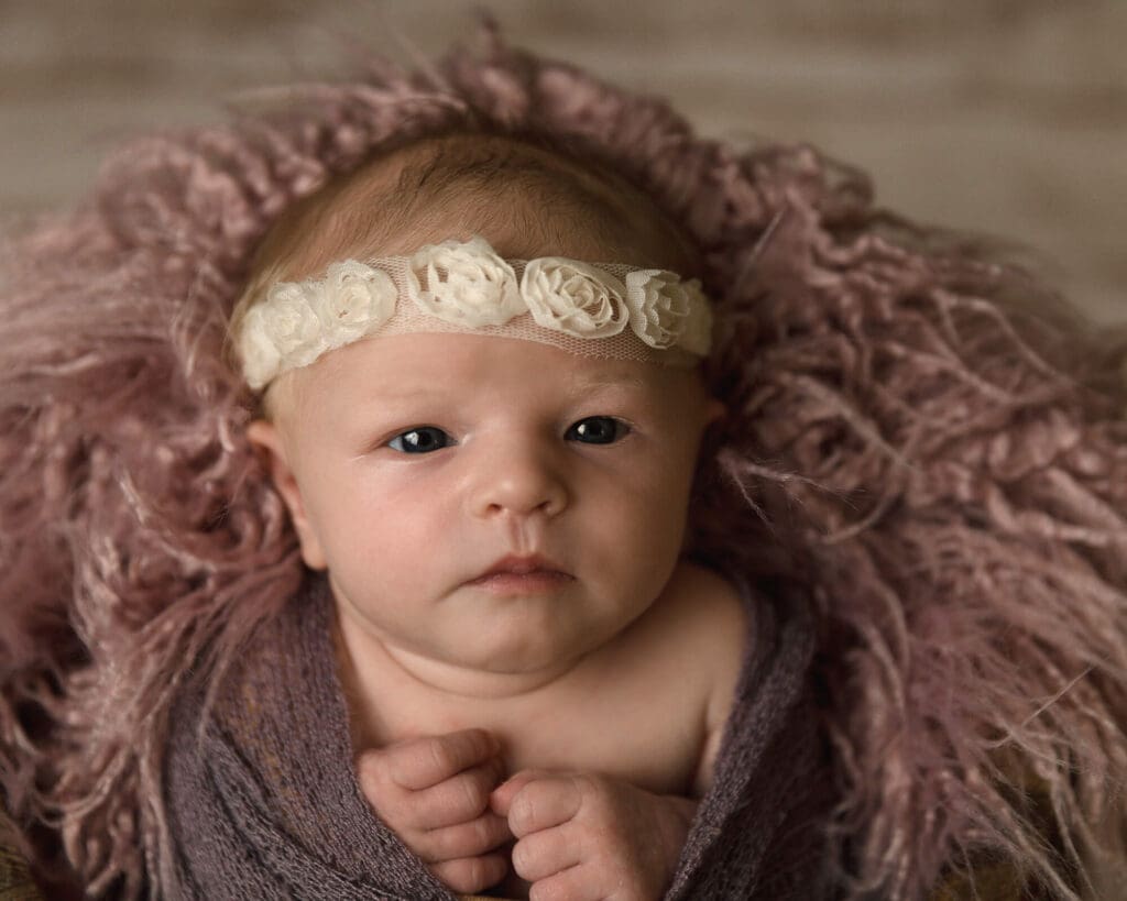 Newborn girl wrapped in purple wrap and wearing a white lace headband laying wide awake.