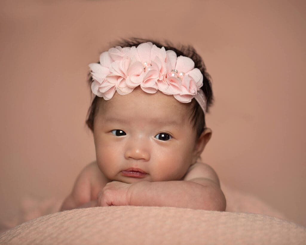 Wide awake newborn girl wearing pink headband resting her chin on her arm. Peach color background.