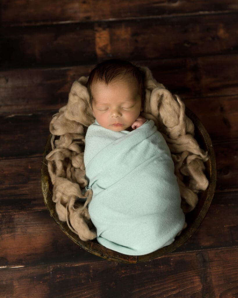 Newborn boy with dark hair wrapped in a light green wrap laying in a bucket filled with tan wool.