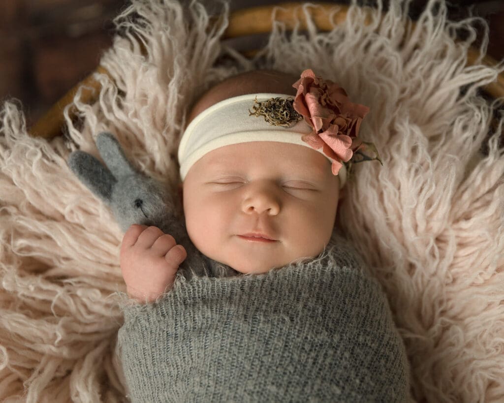 Baby girl wrapped in a gray swaddle, wearing a pink flower headband sleeping on a pink rug as she hold onto a gray wool bunny.