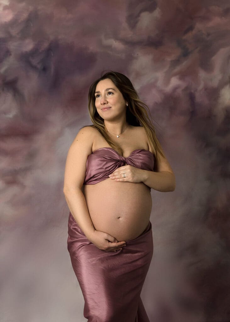 Pregnant woman holding her belly looking away while wrapped in mauve silk fabric.