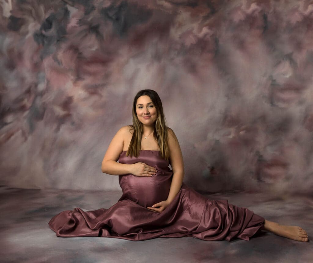 Pregnant woman sitting on the floor holding her belly bump wearing mauve fabric.