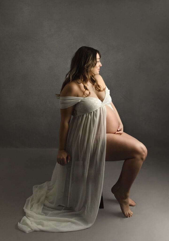 Pregnant woman in a white silky maternity gown sitting holding her belly and looking away.