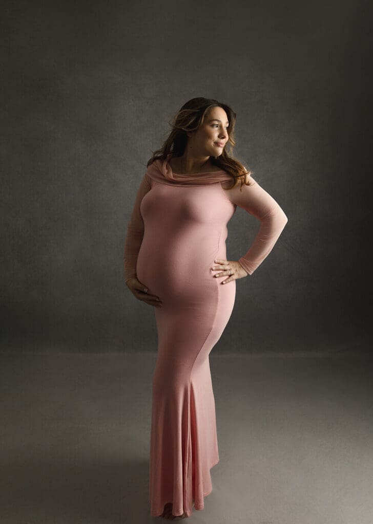 Pregnant woman in a pink maternity dress looking away with one hand on her hip and the other on her belly.